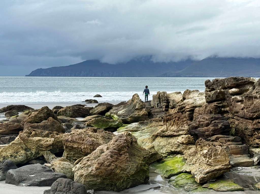 A person stands on rocky beach terrain, gazing out at the ocean under a cloudy sky, with mountains visible in the distance.